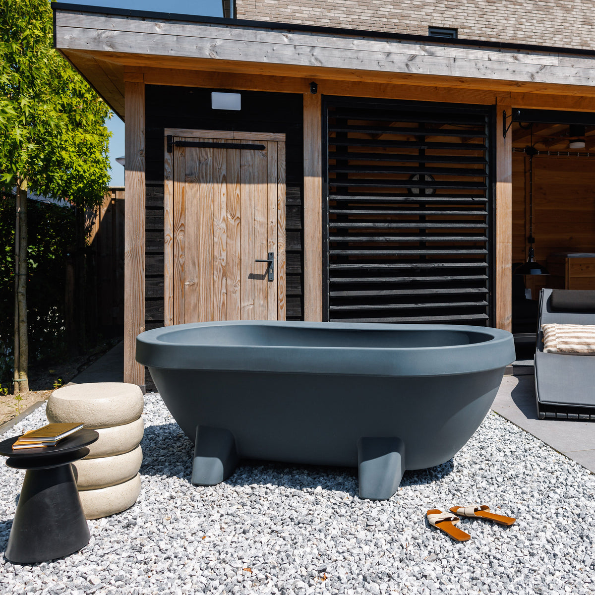 Anthracite Gardentub outdoor bath on white gravel in front of a timber pergola